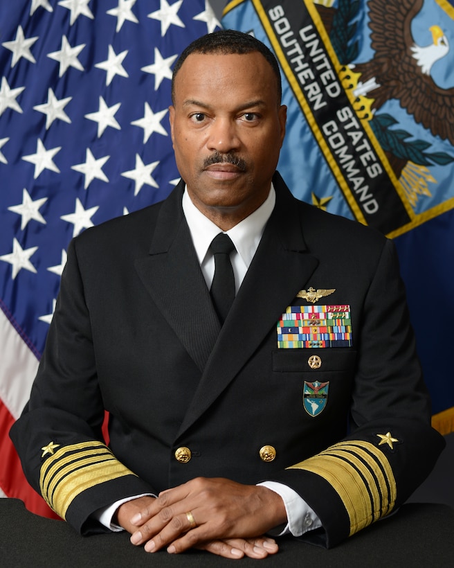 A man in a black navy uniform poses for a photo in front of flags.