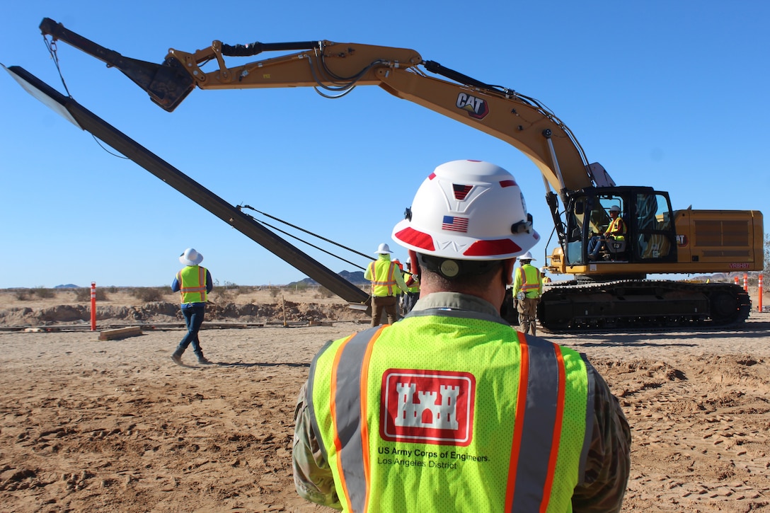 a modified excavator hoists a steel panel off the ground as a U.S. Army Corps of Engineers Soldier watches. The Soldier is wearing a hard hat and a safety vest over his camouflage uniform.