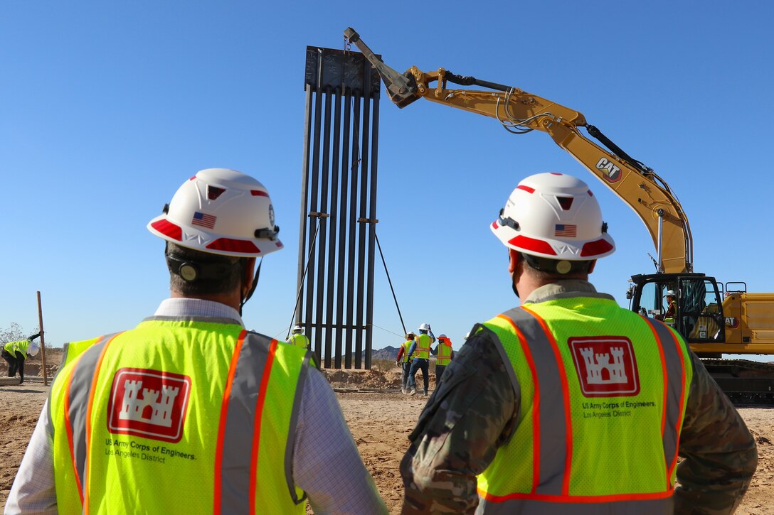 a steel panel is lifted in to position as two members of the U.S. Army Corps of Engineers in hard hats and safety vests look on.