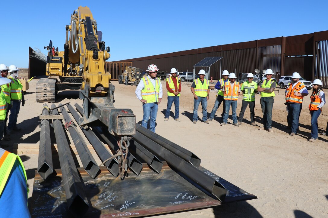 A steel panel is readied to be placed into position as a group of individuals in hard hats and safety vests look on.