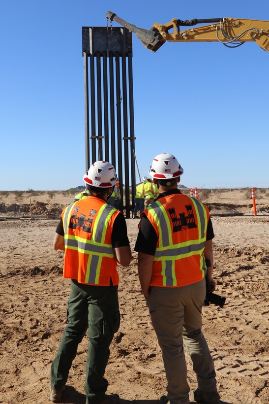 Two U.S. Army Corps of Engineers representatives in hard hats and protective vests look on as a steel panel is placed in along the U.S. southern border.