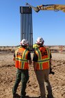 Two U.S. Army Corps of Engineers representatives in hard hats and protective vests look on as a steel panel is placed in along the U.S. southern border.