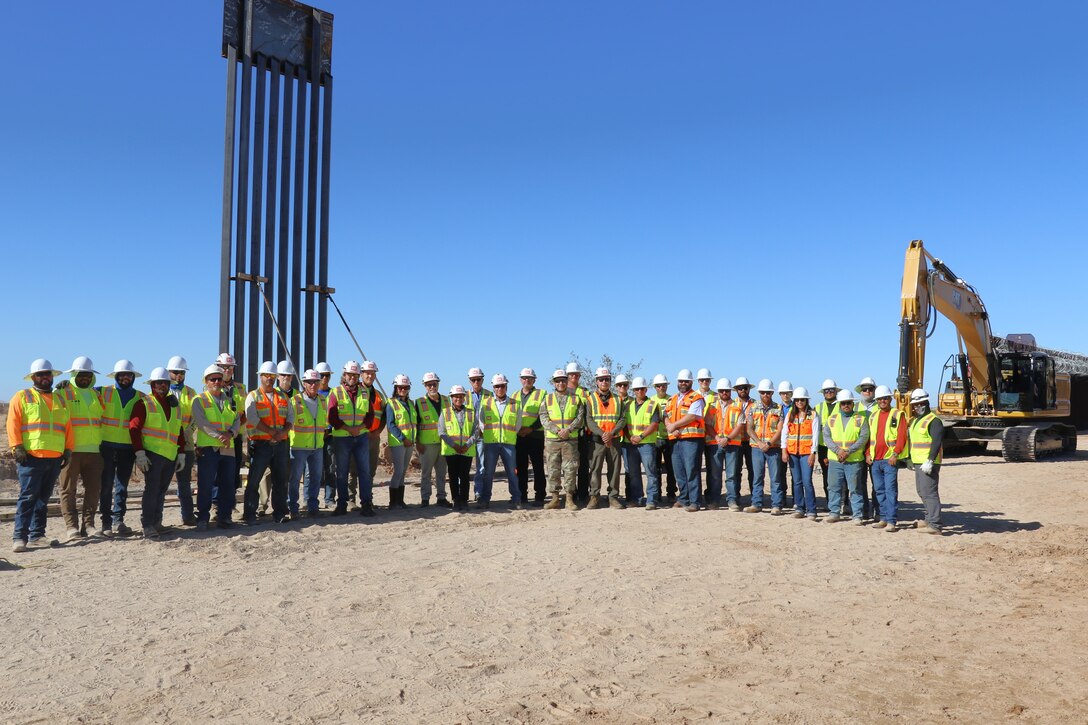 A large group of construction workers and representatives from the U.S. Army Corps of Engineers stands in front of a newly placed steel panel along the U.S. southern border.