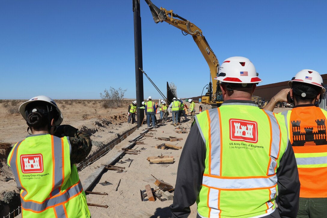 A crane places a large steel barrier into place as three members of the U.S. Army Corps of Engineers wearing hard hats and protective vests observe the action.