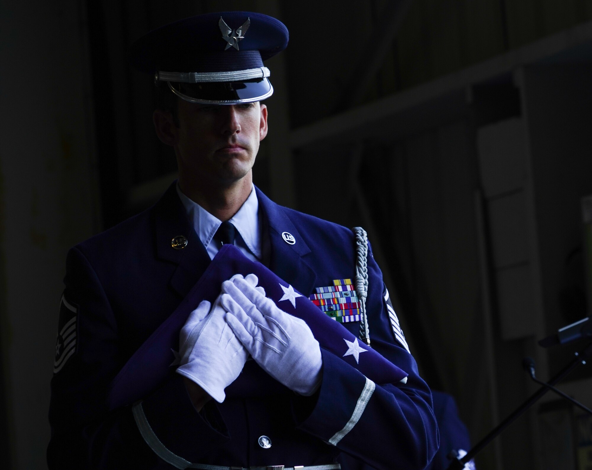 A U.S. Air Force honor guard detail holds a folded flag clutched tight to his chest. A dramatic 4-to-1 lighting ratio is displayed across the left side of his face fixed in front of a dark black background.