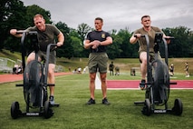 U.S. Marine Corps Gunnery Sgt. Joseph Stich, a force fitness instructor trainer with Training Command, center, instructs Sgt Maj. Michael Croft, command senior enlisted leader with Headquarters and Service Battalion, Headquarters Marine Corps Henderson Hall, left, and a student with the National Capital Regions Staff Sergeant Transition Symposium through an exercise fan bike workout during a physical training event at Butler Stadium on Marine Corps Base Quantico, Virginia, Aug. 6, 2025. The Fiscal Year 25 NCR Staff Sergeant Transition Symposium is a five-day dynamic professional military education course comprised of FY25 Staff Sergeant selectees from diverse Military Occupational Specialty backgrounds and commands, aiming to shape Marines into ethical, operationally grounded SNCOs. (U.S. Marine Corps photo by Lance Cpl. Braydon Rogers)