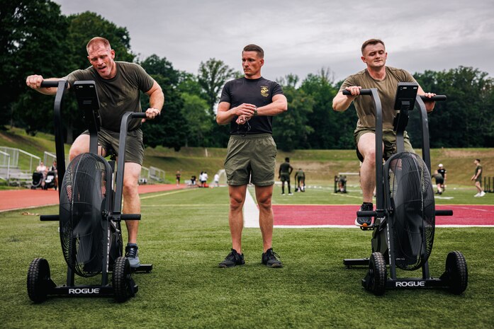 U.S. Marine Corps Gunnery Sgt. Joseph Stich, a force fitness instructor trainer with Training Command, center, instructs Sgt Maj. Michael Croft, command senior enlisted leader with Headquarters and Service Battalion, Headquarters Marine Corps Henderson Hall, left, and a student with the National Capital Regions Staff Sergeant Transition Symposium through an exercise fan bike workout during a physical training event at Butler Stadium on Marine Corps Base Quantico, Virginia, Aug. 6, 2025. The Fiscal Year 25 NCR Staff Sergeant Transition Symposium is a five-day dynamic professional military education course comprised of FY25 Staff Sergeant selectees from diverse Military Occupational Specialty backgrounds and commands, aiming to shape Marines into ethical, operationally grounded SNCOs. (U.S. Marine Corps photo by Lance Cpl. Braydon Rogers)