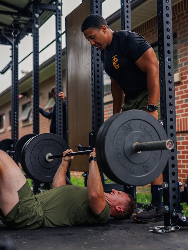 U.S. Marine Corps Staff Sgt. Donald Baker, a force fitness instructor trainer with Training Command, spots a student with the National Capital Regions Staff Sergeant Transition Symposium during a floor bench press while conducting a physical training event at Butler Stadium on Marine Corps Base Quantico, Virginia, Aug. 6, 2025. The Fiscal Year 25 NCR Staff Sergeant Transition Symposium is a five-day dynamic professional military education course comprised of FY25 Staff Sergeant selectees from diverse Military Occupational Specialty backgrounds and commands, aiming to shape Marines into ethical, operationally grounded SNCOs. (U.S. Marine Corps photo by Lance Cpl. Braydon Rogers)