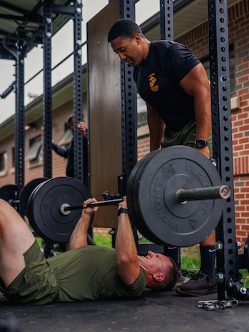 U.S. Marine Corps Staff Sgt. Donald Baker, a force fitness instructor trainer with Training Command, spots a student with the National Capital Regions Staff Sergeant Transition Symposium during a floor bench press while conducting a physical training event at Butler Stadium on Marine Corps Base Quantico, Virginia, Aug. 6, 2025. The Fiscal Year 25 NCR Staff Sergeant Transition Symposium is a five-day dynamic professional military education course comprised of FY25 Staff Sergeant selectees from diverse Military Occupational Specialty backgrounds and commands, aiming to shape Marines into ethical, operationally grounded SNCOs. (U.S. Marine Corps photo by Lance Cpl. Braydon Rogers)