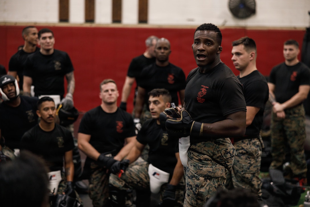 U.S. Marine Corps Sgt. Arkel Brown, right, a Martial Arts Instructor Trainer and mortarman, with the Martial Arts and Fitness Center of Excellence, The Basic School, speaks to Marines attending the Marine Corps Martial Arts 25th anniversary symposium hosted by the MAFCE pose for a group photo on Marine Corps Base Quantico, Virginia, Oct. 9, 2025. The goal of this symposium was to provide the Fleet Marine Force with instructions and practical application on the new tan-black belt syllabus and the new free sparring events while integrating guided discussions fostering character development and recounting the storied tradition of the Marine Corps Martial Arts Program. (U.S. Marine Corps photo by Cpl. Joshua Barker)