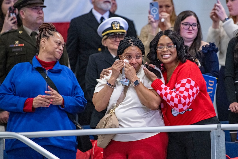 Three civilians watch an event and emote while standing in front of a crowd – one wiping away tears and two others smiling.