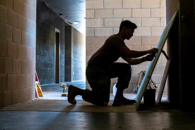 A kneeling sailor, shown in silhouette, handles a piece of drywall resting against a wall in a hallway.