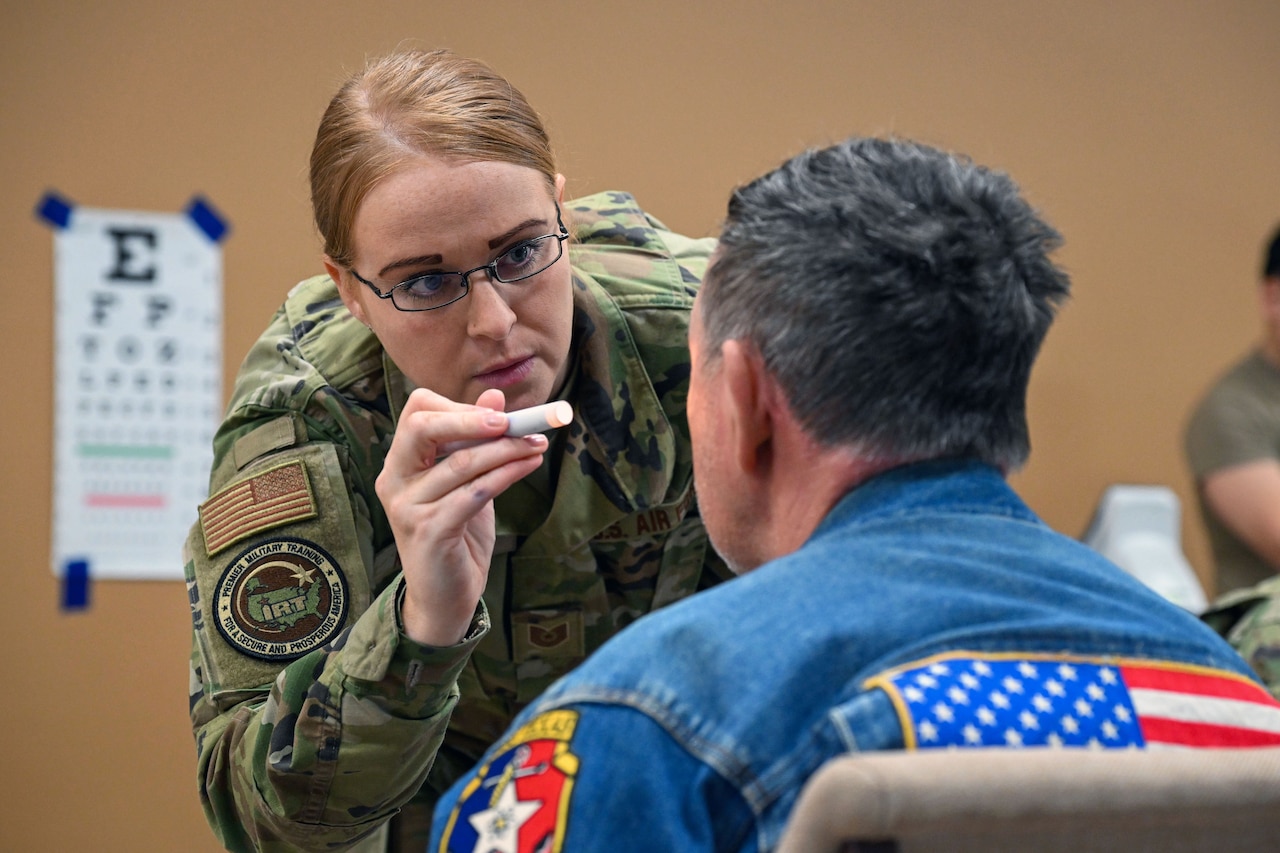 An airman shines a hand-held light into the left eye of a civilian sitting in a chair facing an eye chart taped to a wall.