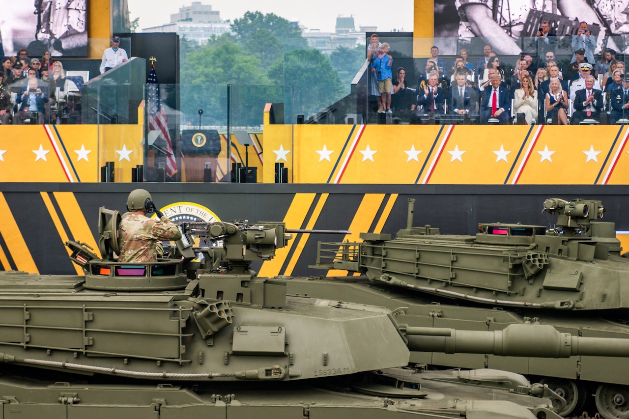 A soldier shown from behind while standing in the turret of a tank salutes the president, who returns the salute while sitting in elevated reviewing stand with other dignitaries.