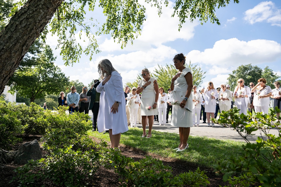 People in white dresses, some with bowed heads and one hand over their hearts, hold white flowers while standing near a tree in a cemetery.