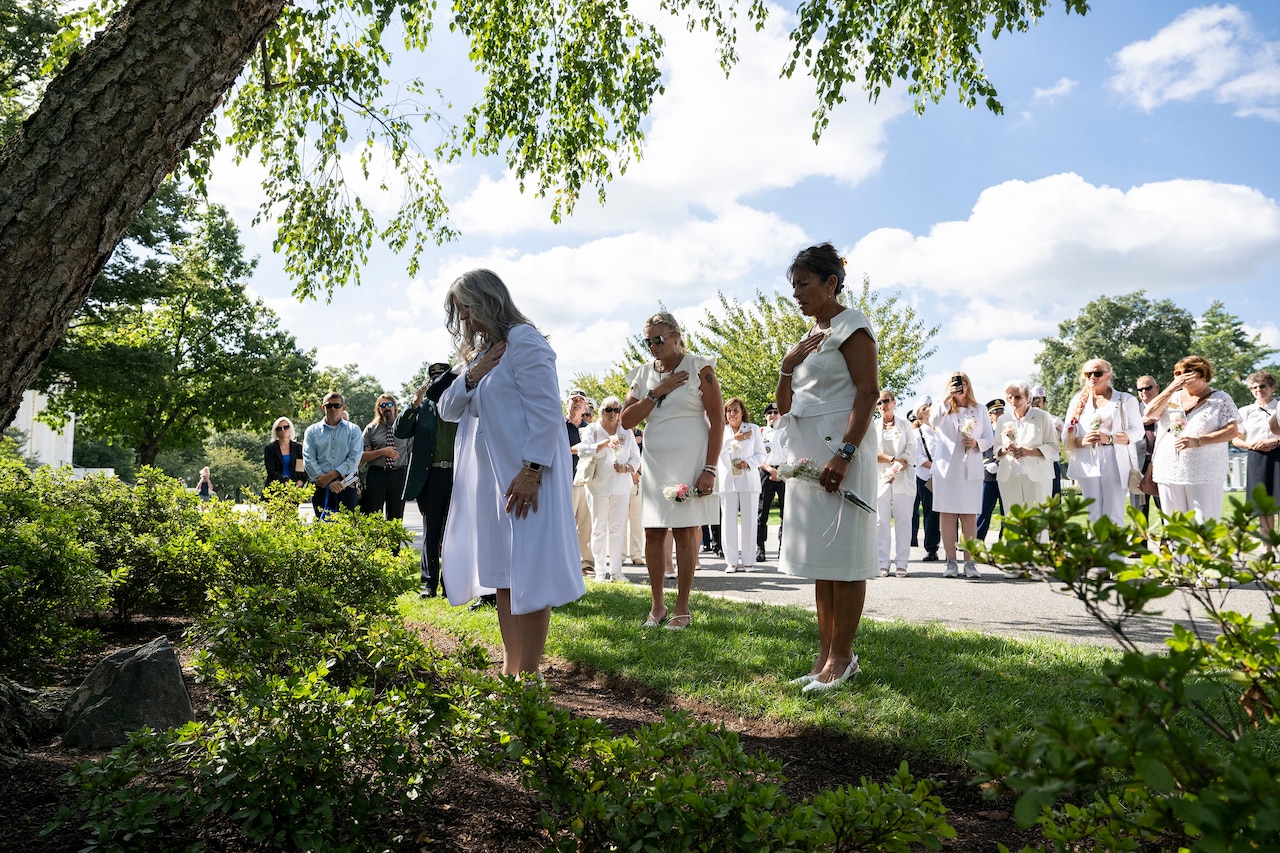People in white dresses, some with bowed heads and one hand over their hearts, hold white flowers while standing near a tree in a cemetery.