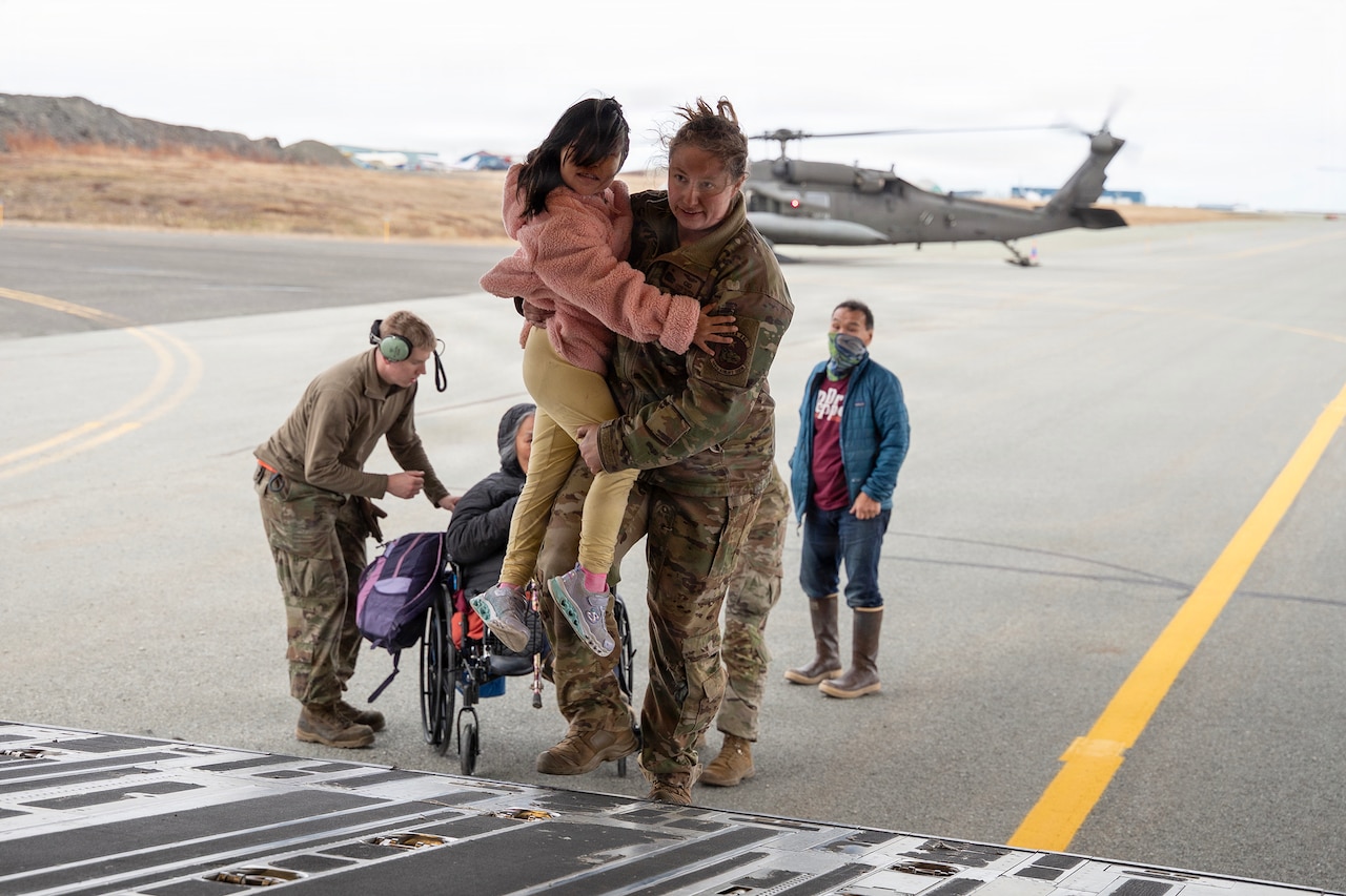 An airman carries up a child up a metal ramp into a parked aircraft as another service member helps civilians on the flight line.