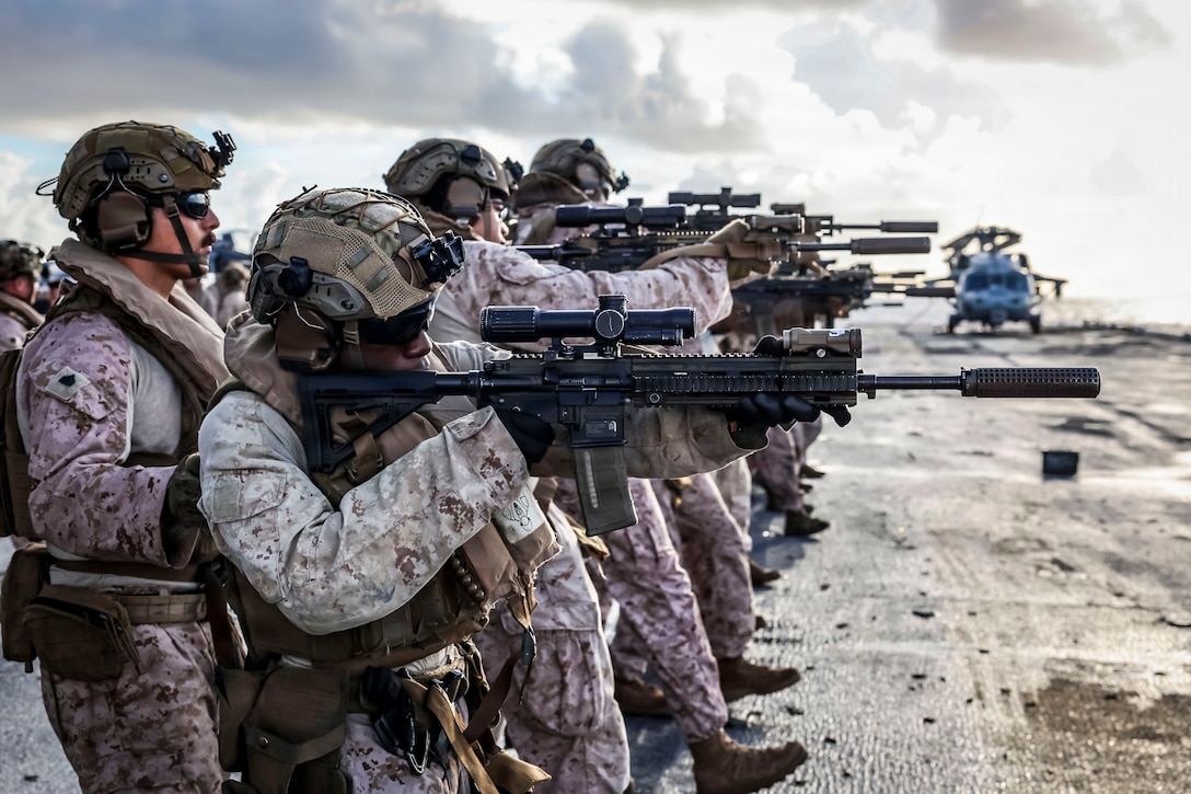 U.S. military members in camouflage uniforms and helmets point rifles.