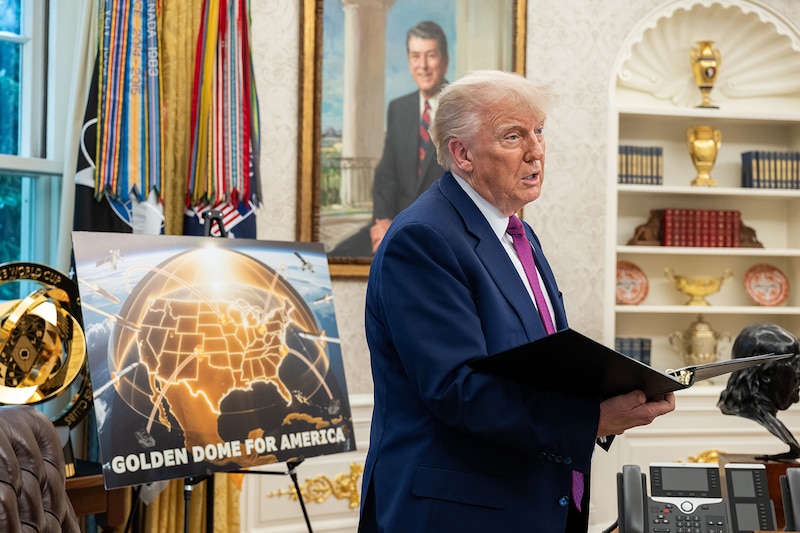 The president stands and holds a folder while speaking in an office with a "Golden Dome for America" poster displayed behind him.