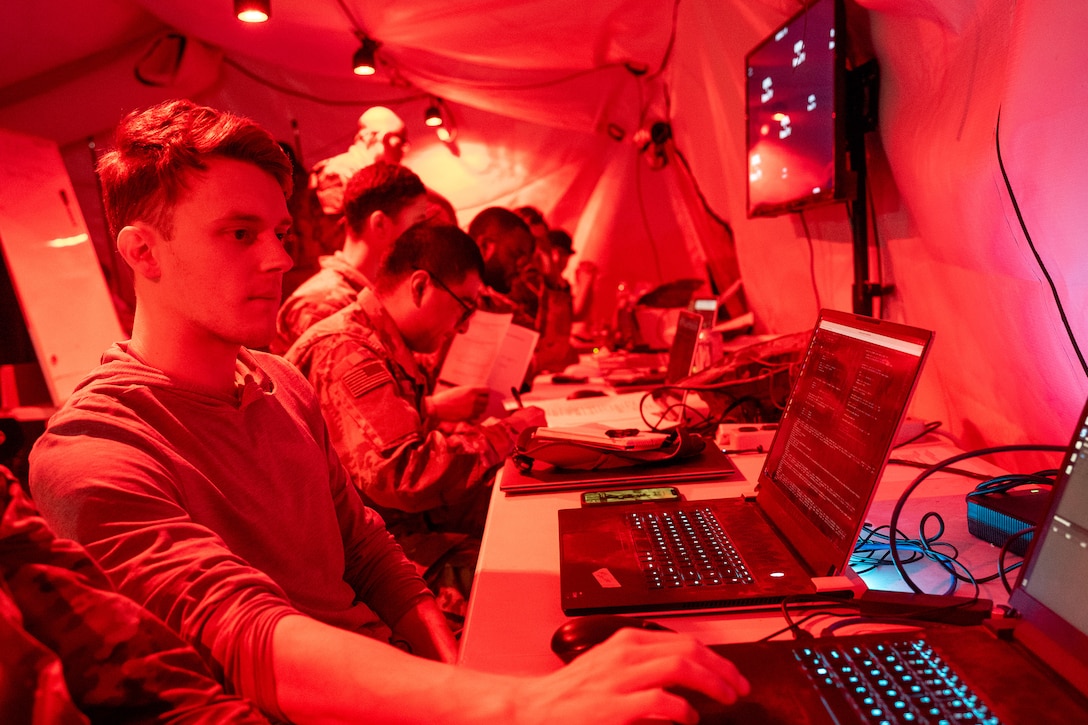 Civilians and service members sit in front of laptops at a table in a tent-like space lit by red light.