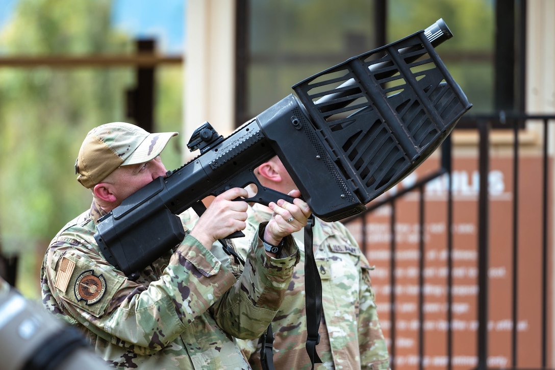 An airman aims a large, black weapon upwards outside a building.