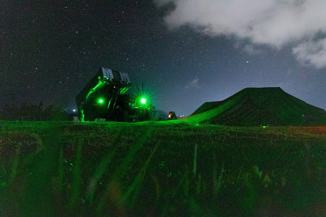 A weapon system sits in a dark field illuminated by green light with a mountain in the background.
