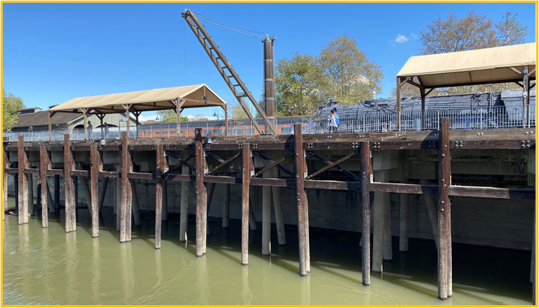 river water site in the foreground and a raised wooden platform where a train is stationed and people are walking along the water.