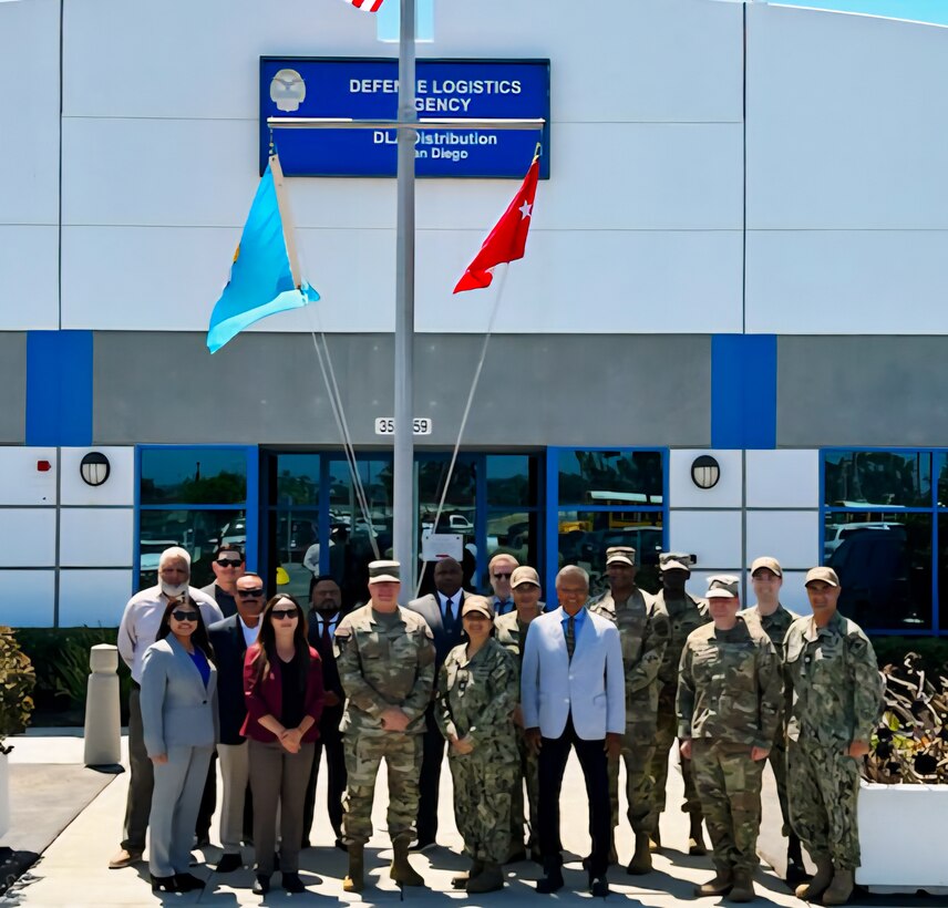 A group of 17 people, eight of them wearing camouflage military uniforms, stand outside on a sunny day, underneath a flagpole in front of a building with a sign on it that says 'Defense Logistics Agency Distribution San Diego'.