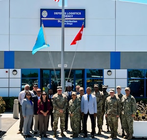 A group of 17 people, eight of them wearing camouflage military uniforms, stand outside on a sunny day, underneath a flagpole in front of a building with a sign on it that says 'Defense Logistics Agency Distribution San Diego'.
