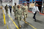A group of people, four of them wearing camouflage military uniforms, walk through a large warehouse.