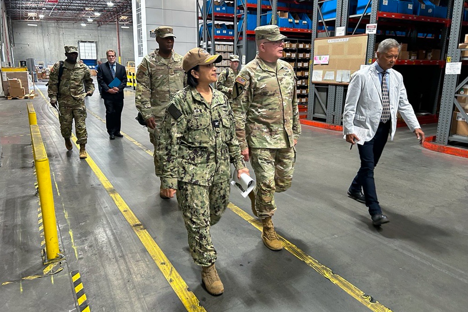 A group of people, four of them wearing camouflage military uniforms, walk through a large warehouse.