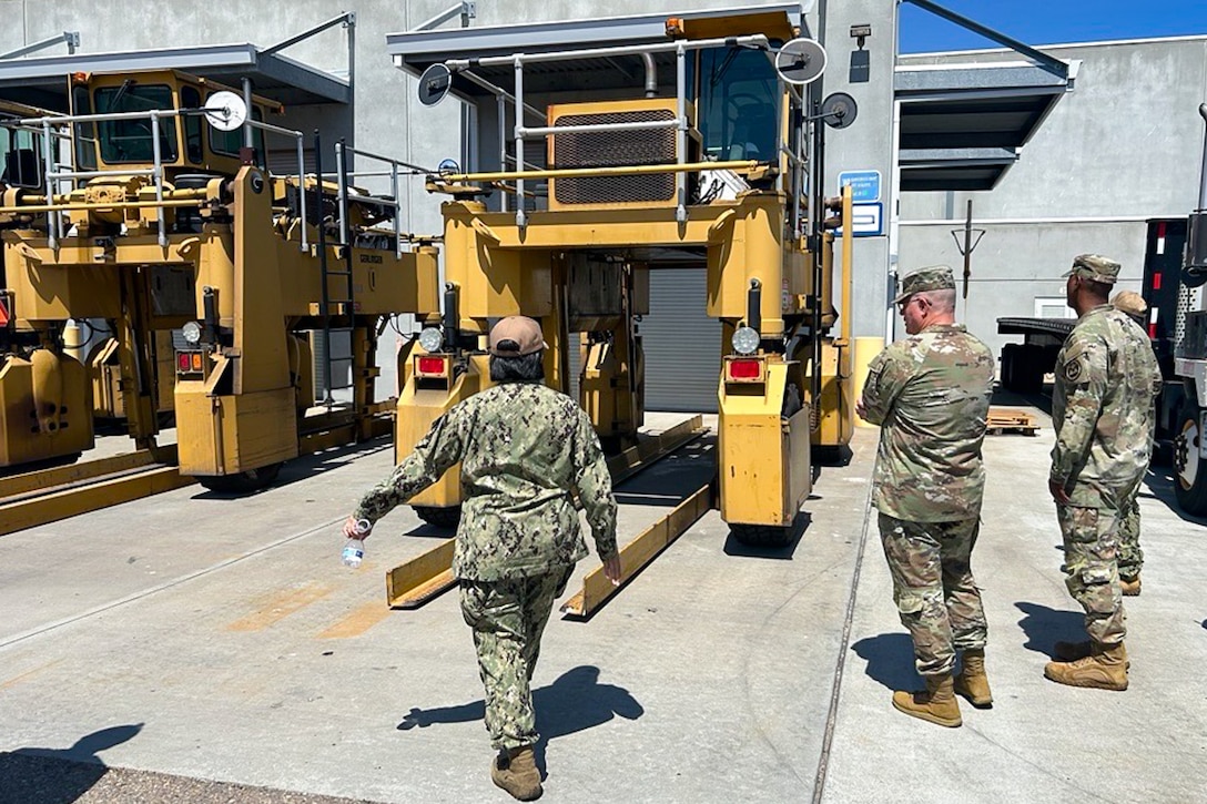 A group of people wearing camouflage military uniforms stand outside looking at large machinery.