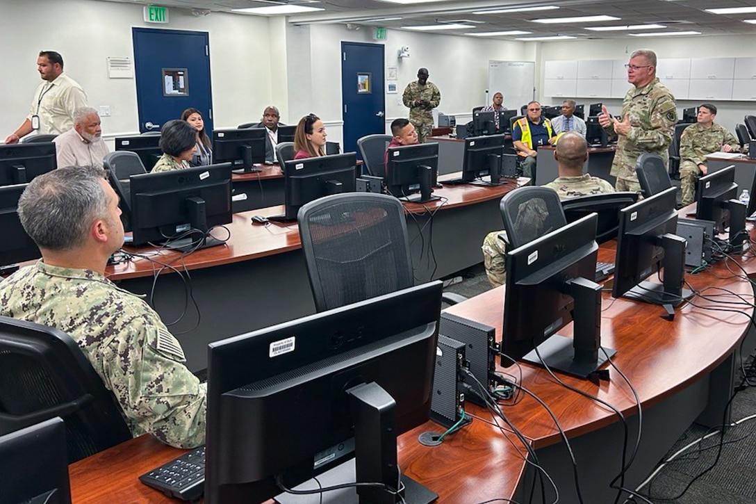 A man wearing a camouflage military uniform speaks to a group of people who are seated in a room at multiple long tables with computers on them.