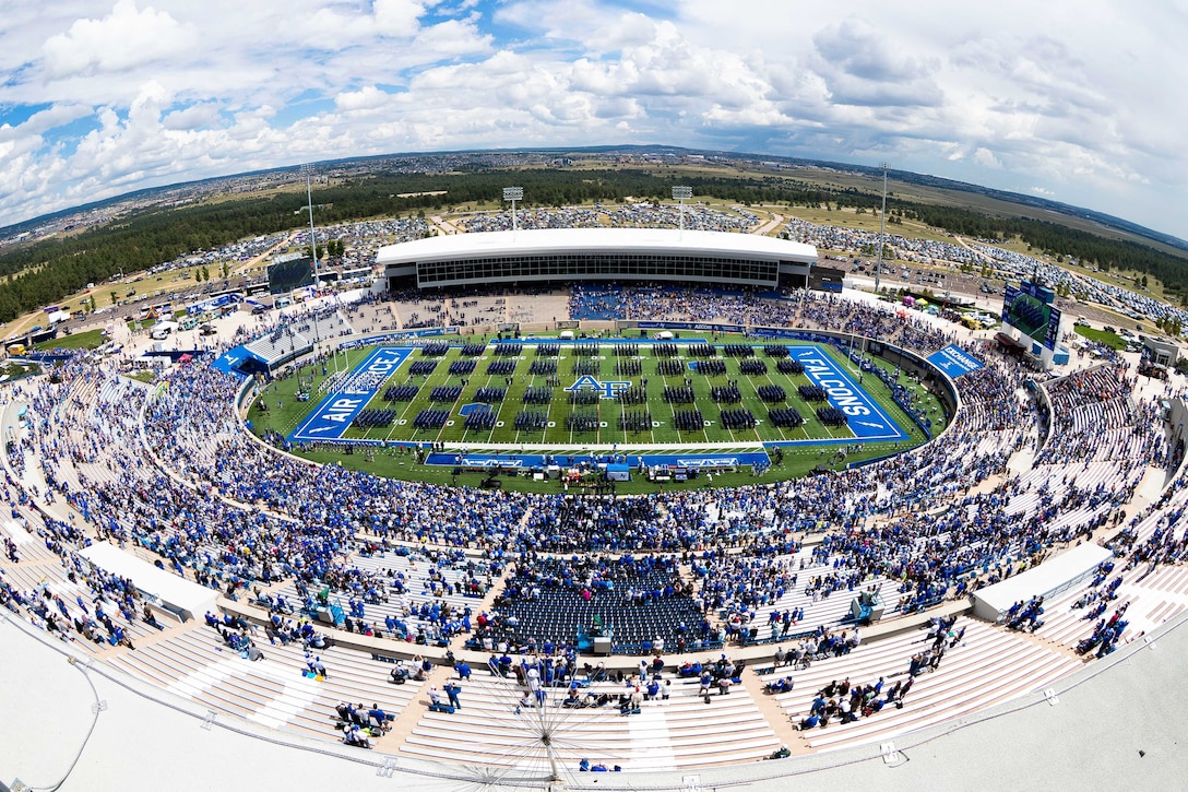 Dozens of U.S. Air Force Academy cadets march on a field as hundreds of fans watch from the bleachers in a stadium as seen through a fisheye lens.