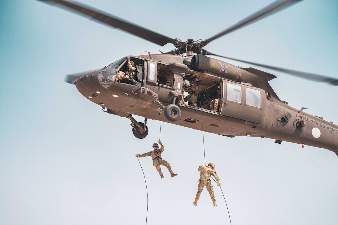 Two soldiers rappel from an airborne helicopter against a light blue sky.