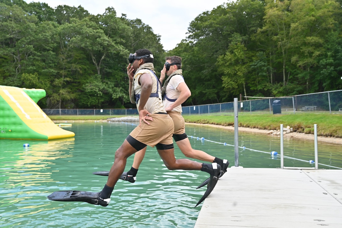 Two sailors wearing goggles and fins jump into a body of water near a wooded area.