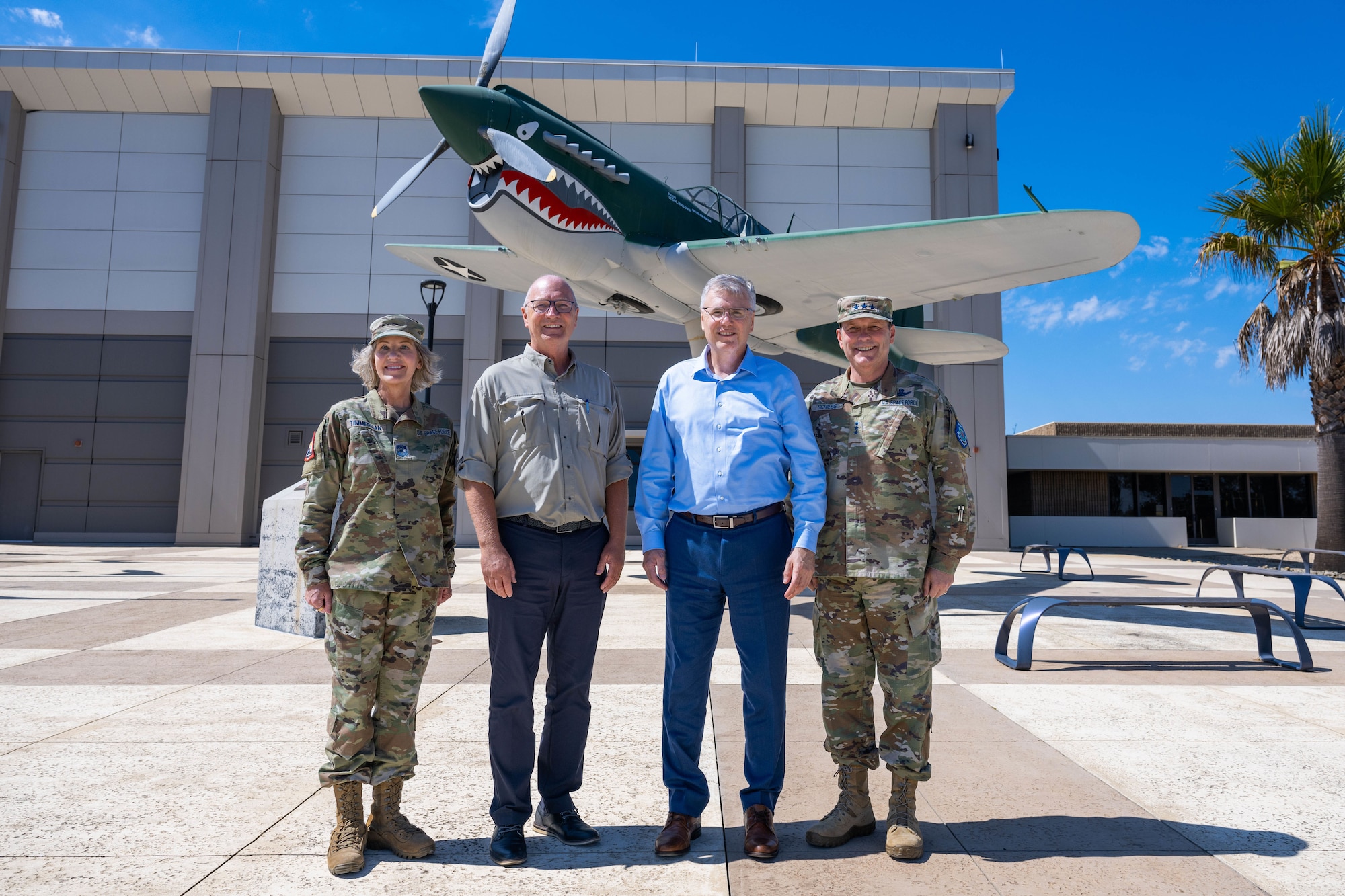 Four people stand shoulder-to-shoulder for a group photo outside a building and in front of a static display of a P-40 aircraft.