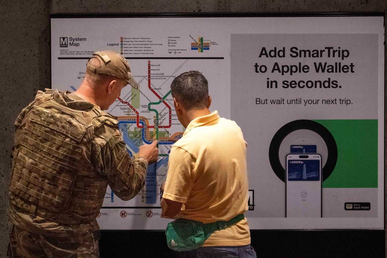 An airman helps a person in civilian clothing navigate a train map with different colors on it on a sign indoors.
