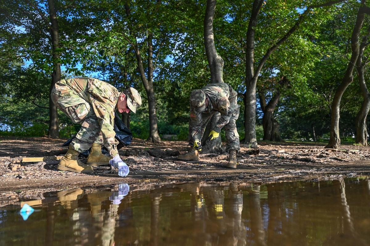 Two men dressed in camouflage military uniforms bend down to pick up trash and debris along a waterway. Trees can be seen in the background.