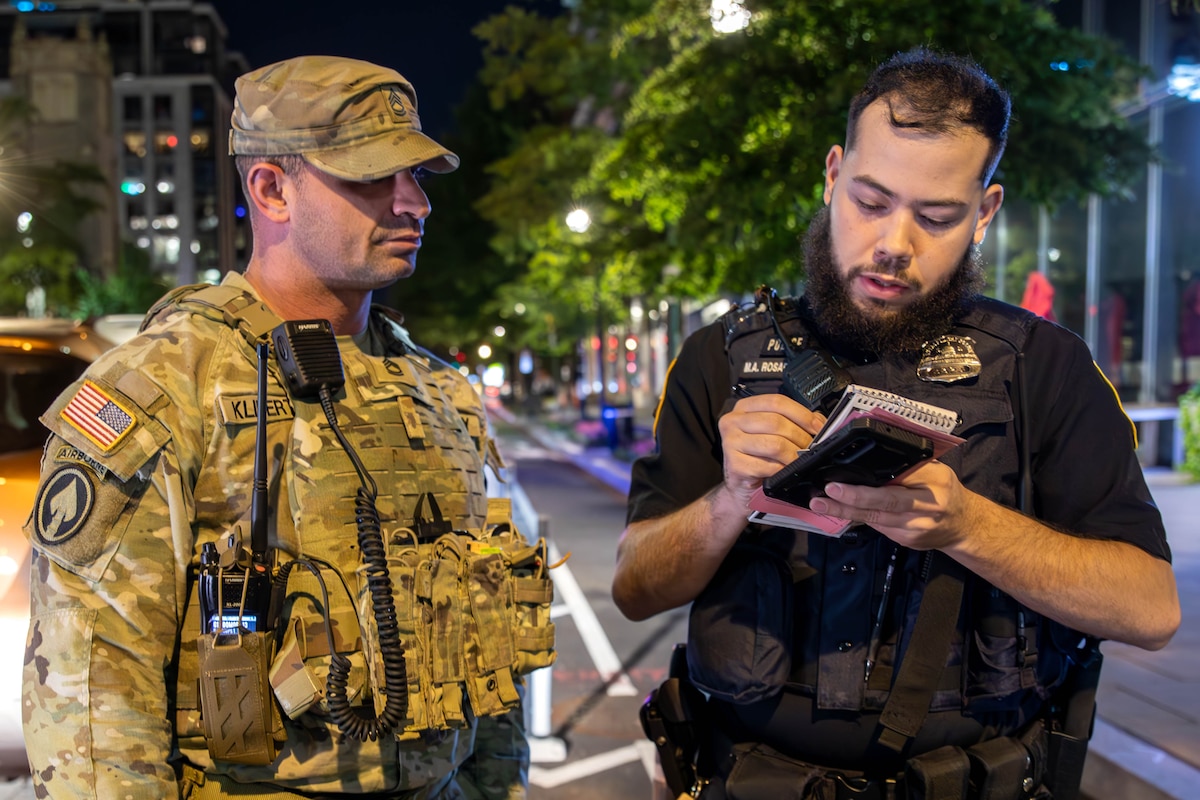 A man to the left, who is dressed in a camouflage military uniform, speaks to a man dressed in a black police uniform, writing on a notepad.