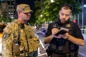 A man to the left, who is dressed in a camouflage military uniform, speaks to a man dressed in a black police uniform, writing on a notepad.