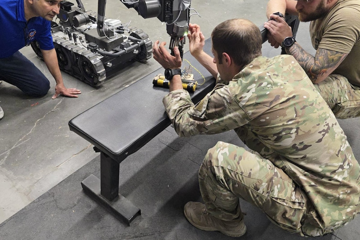 A person wearing a camouflage military uniform squats in front of a robot to look at a notional explosive device as another person dressed in a camouflage military uniform and a person in civilian attire watch.