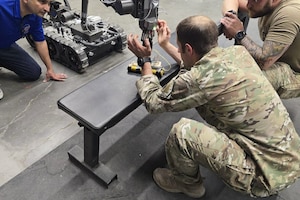 A person wearing a camouflage military uniform squats in front of a robot to look at a notional explosive device as another person dressed in a camouflage military uniform and a person in civilian attire watch.