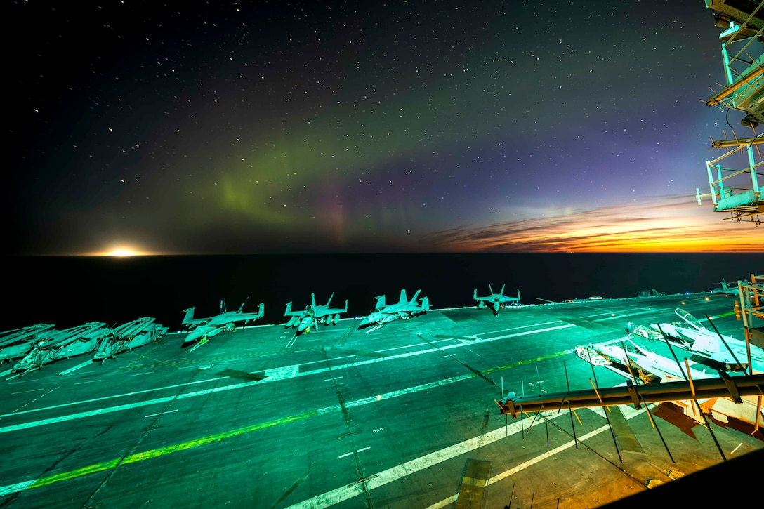 Green and blue lights are shown in a night sky from a ship's deck at sea, which has parked aircraft on it and is bathed in green light.