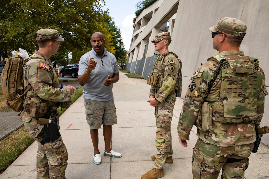 Three guardsmen stand and speak with a person in a grey shirt while on a sidewalk next to a tan building on a sunny day.