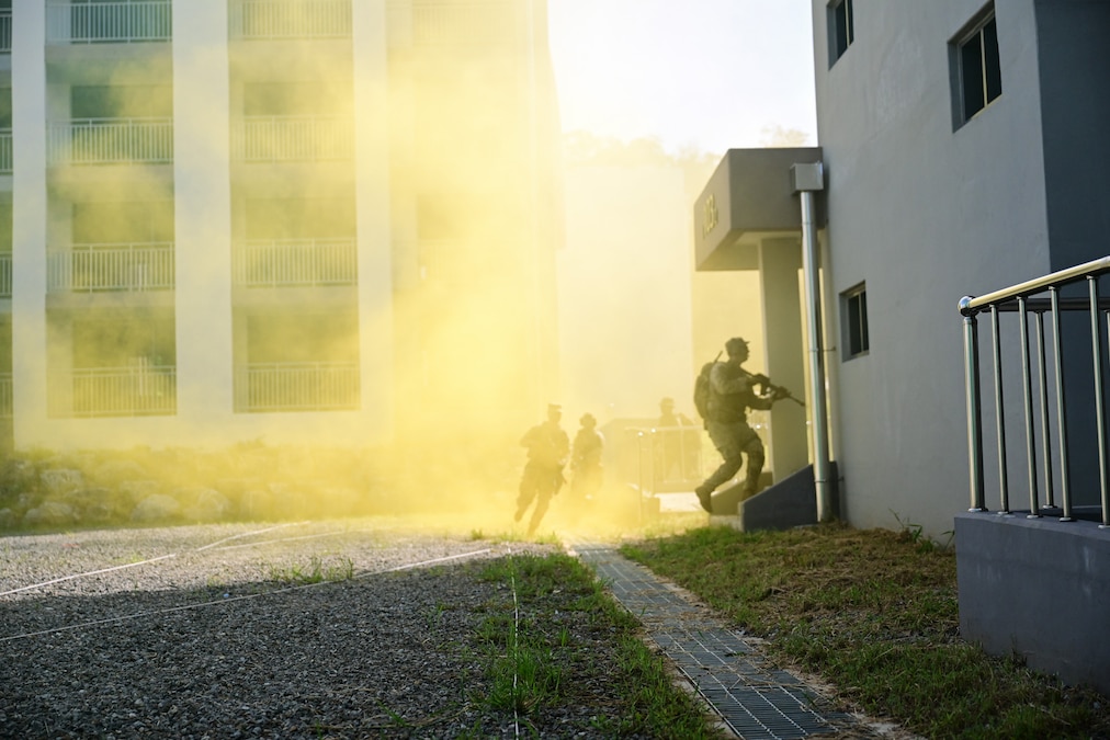 Four soldiers in tactical gear hold service weapons and run through yellow smoke between two grey buildings during the day.