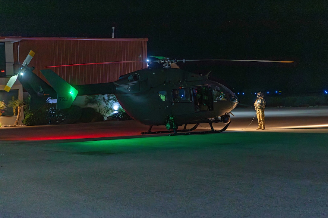 A soldier stands in front of a helicopter parked on a runway at night.