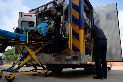 EMS worker stands next to the ambulance as patient is loaded onto the stretcher. Another EMS personnel loads patient onto stretcher.