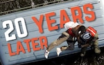 The words 20 Years Later are shown on the top of a bus. In the foreground, a Coast Guard rescue swimmer is hoisting a survivor during Hurricane Katrina.