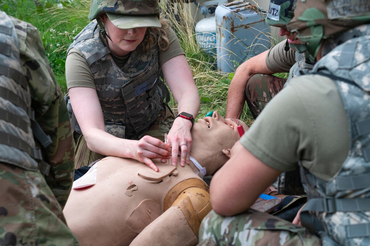 A woman wearing a military camouflage uniform and helmet places a needle in a simulated casualty mannequin while other people in similar attire observe.